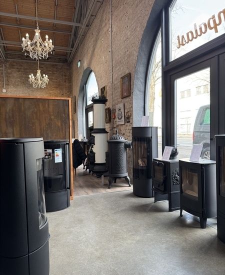 Ein gemütlicher Ausstellungsraum mit verschiedenen modernen und traditionellen Öfen, geschmückt mit einem Kronleuchter und warmen Backsteinwänden.