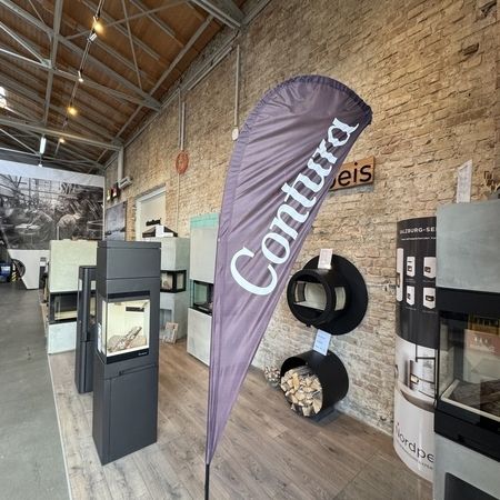 Ein Banner mit der Marke Contura steht in einem modernen Ausstellungsraum, in dem verschiedene stilvolle Holzöfen vor einer Backsteinwand ausgestellt sind.