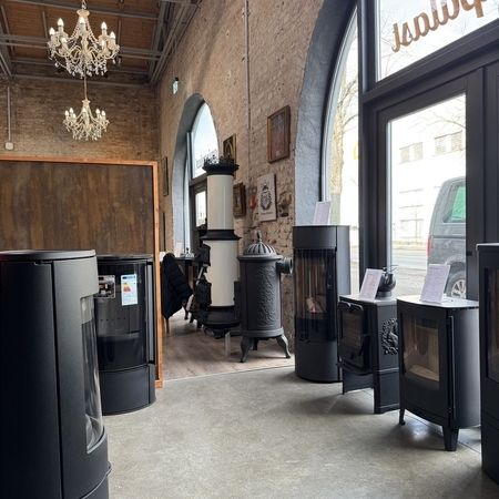 Ein gemütlicher Ausstellungsraum, in dem verschiedene moderne und alte Holzöfen ausgestellt sind, mit eleganten Kronleuchtern und großen Fenstern in einem rustikalen Ambiente.