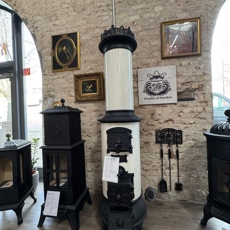 Ein Kaminofen im Vintage-Stil mit weißem und schwarzem Design steht in einem gemütlichen Ausstellungsraum, umgeben von gerahmten Kunstwerken und Kaminzubehör.