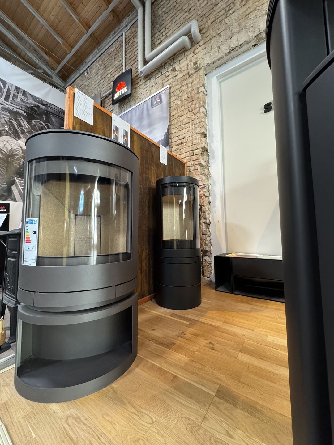 Zwei moderne Holzöfen in einem Ausstellungsraum mit schlichtem schwarzem Design und Glasfronten, die auf einem hellen Holzboden stehen.