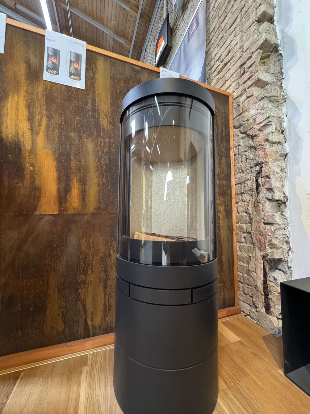 Moderner schwarzer Holzofen mit Glasfront, ausgestellt auf einem Holzboden vor einer strukturierten Ziegelwand und einem goldenen Hintergrund.