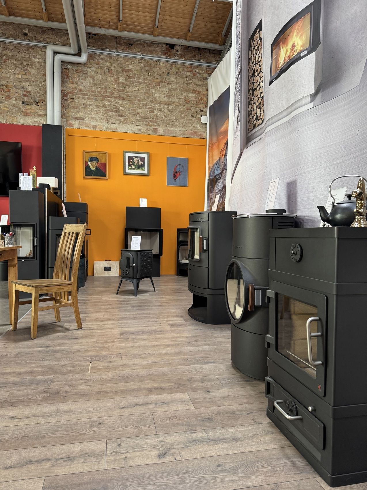 Ein gemütlicher Ausstellungsraum mit verschiedenen modernen schwarzen Öfen an einer farbenfrohen Wand, mit einem Holzstuhl und gerahmter Kunst.
