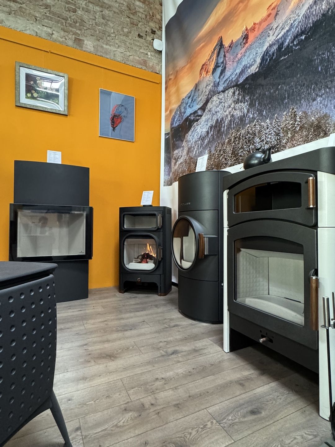 Ein warmes Interieur mit verschiedenen modernen Öfen vor einer orangefarbenen Wand und einer malerischen Berglandschaft im Hintergrund.