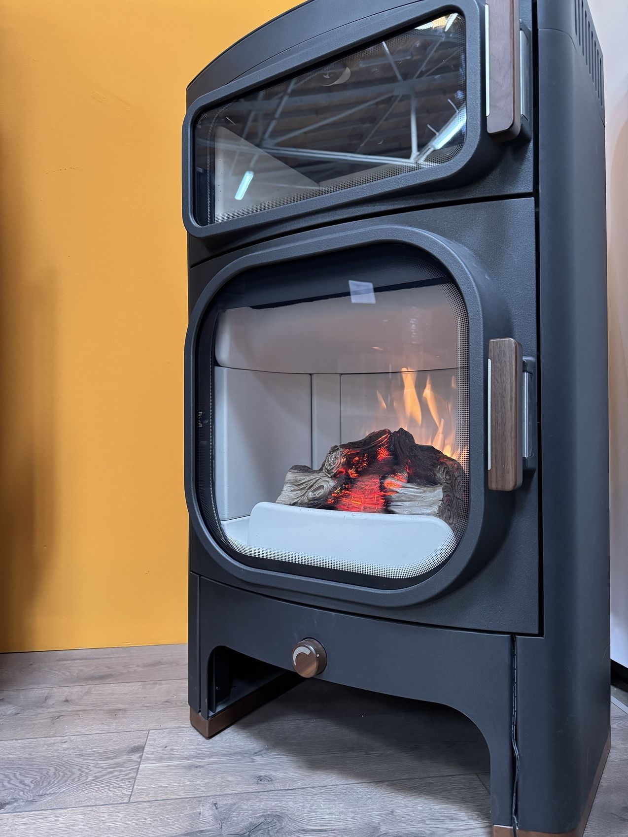 Ein moderner schwarzer Kamin mit Glastüren, in dem glühende Holzscheite und flackernde Flammen zu sehen sind, vor einer orangefarbenen Wand und einem Holzboden.