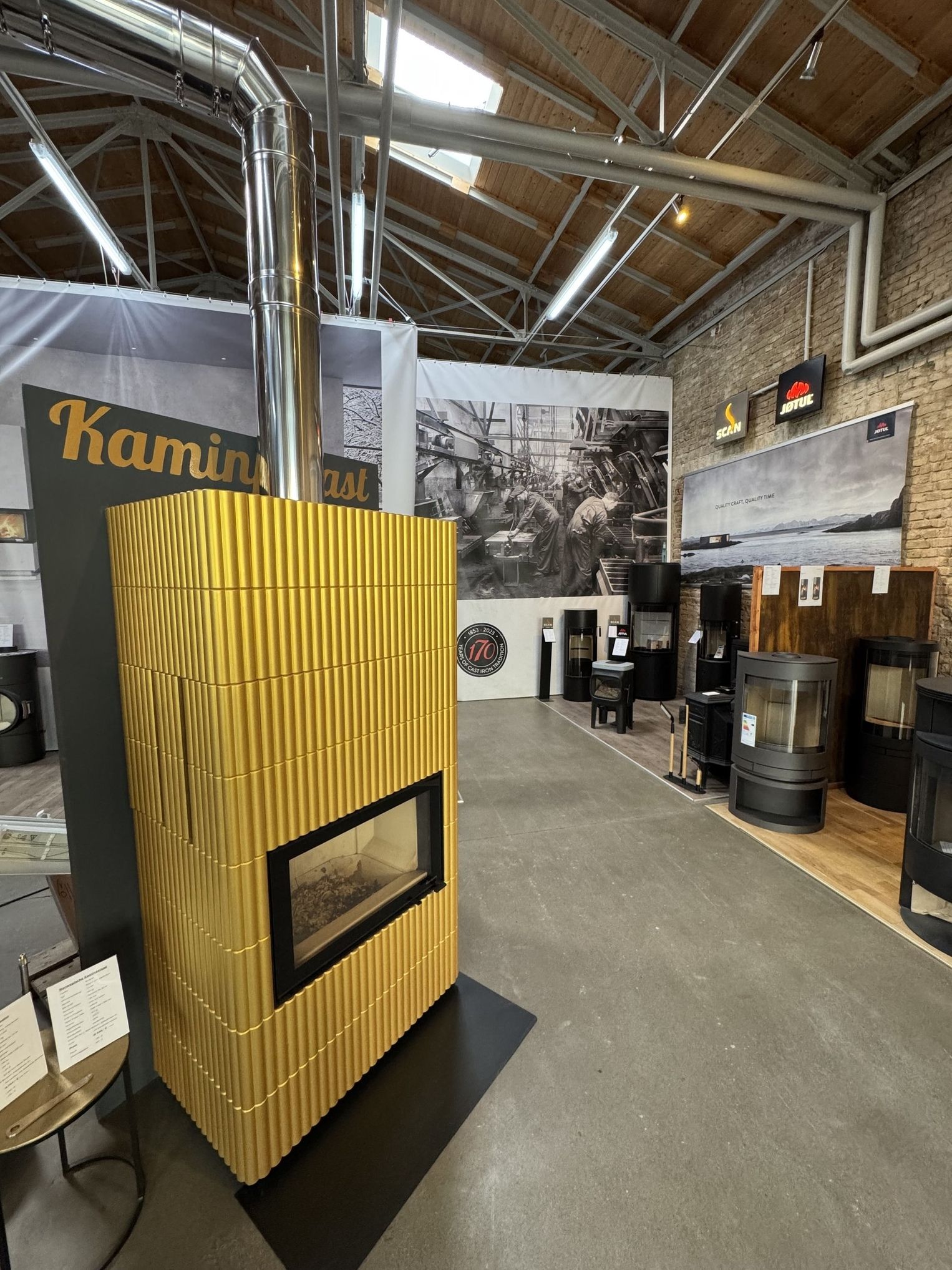 Ein moderner goldener Kamin mit Glasfront und Edelstahlschornstein, der in einem geräumigen, stilvollen Ausstellungsraum mit anderen Designs präsentiert wird.