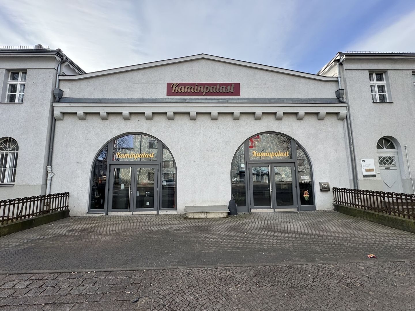 Fassade des Kaminpalastes, eines modernen Gebäudes mit gewölbten Glastüren und einem Schild mit goldener Schrift auf einfachem grauem Hintergrund.