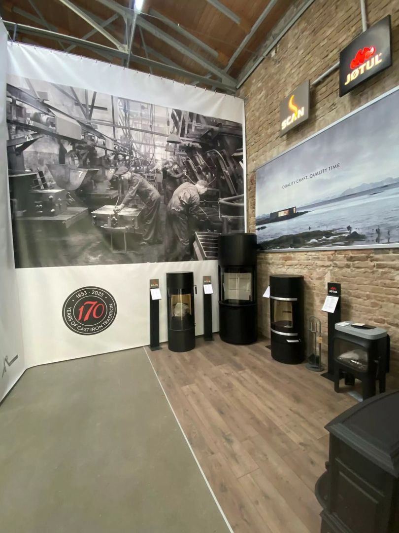 Eine Ausstellung moderner Holzöfen mit einem historischen Schwarz-Weiß-Fotohintergrund, der 170 Jahre Gusseisenhandwerk feiert.